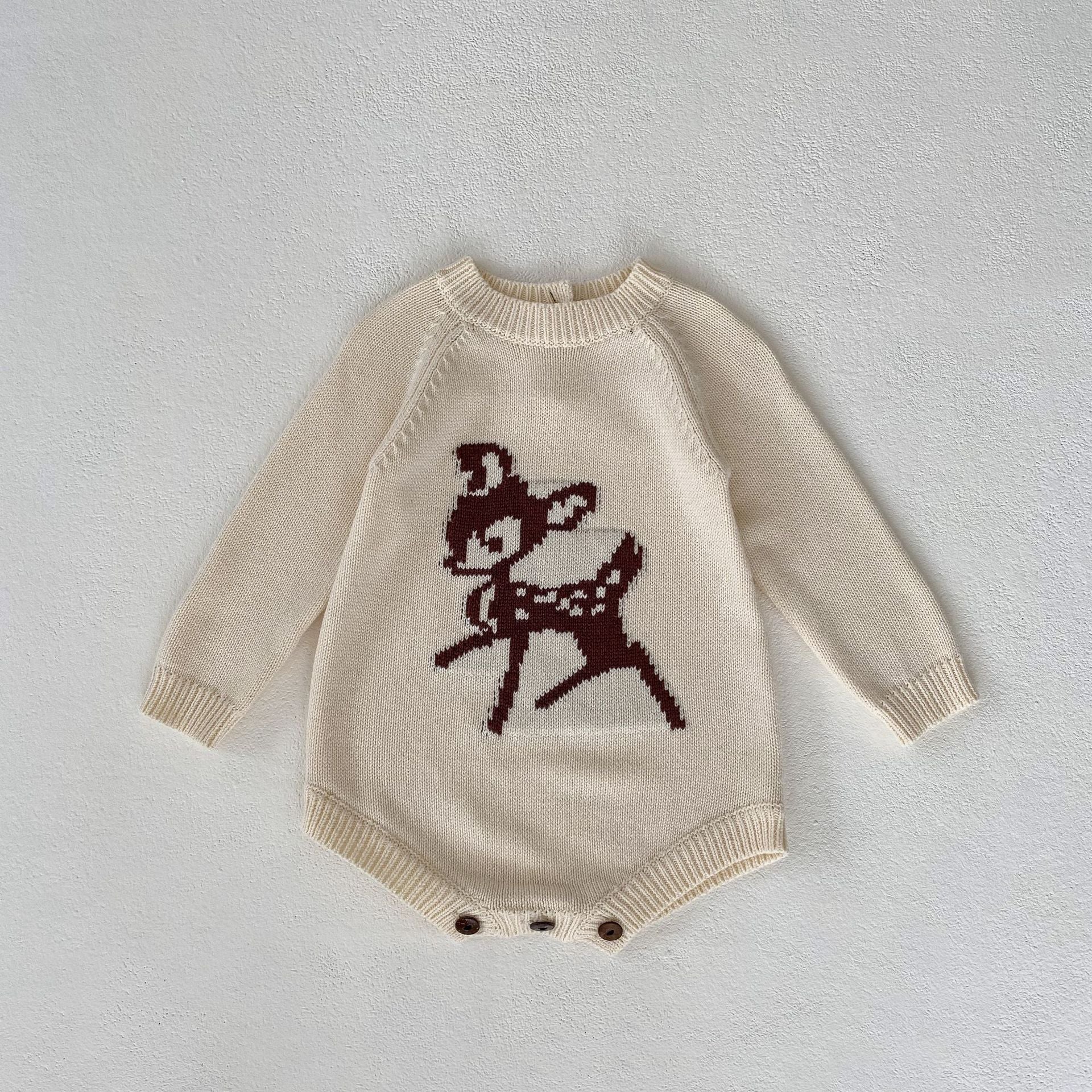 Simple and adorable cream-colored knit romper for babies, featuring a cute brown deer motif on the front, designed for warmth and style during colder seasons. The romper has long sleeves and button closures at the bottom for easy diaper changes. Displayed flat on a neutral textured background, highlighting its soft material and cozy design, making it ideal for fall and winter baby outfits.