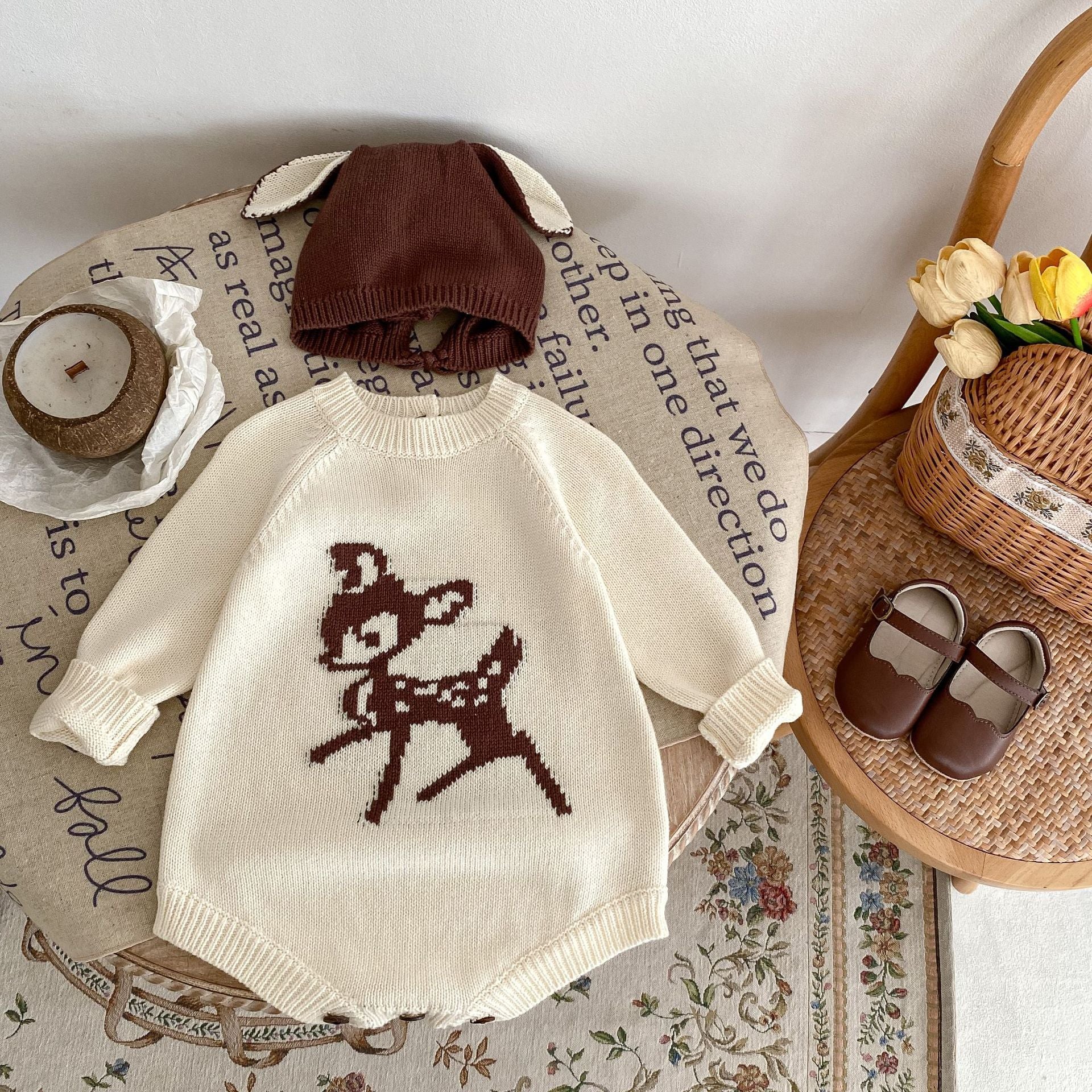 Adorable baby outfit flat lay featuring a cozy beige knit sweater romper with a charming brown deer motif on the front, perfect for fall or winter. Styled with a matching brown beanie hat with cute ear details and a pair of brown baby shoes. The outfit is placed on a round table covered in a fabric with inspirational text, surrounded by autumnal decor, including a wicker basket holding fresh tulips and a coconut candle for a warm and rustic touch. Ideal for capturing cozy, seasonal baby fashion