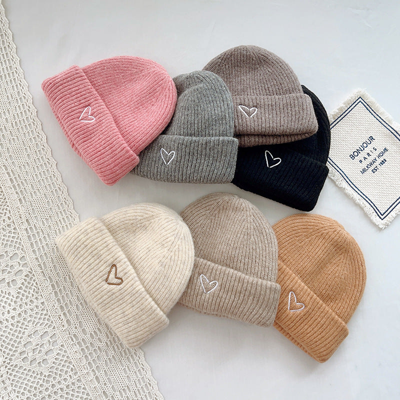 Collection of cozy knitted beanies in a variety of colors, including pink, gray, brown, black, cream, beige, and tan, each featuring a small embroidered heart on the cuff. These soft and stylish hats are ideal for winter wear, adding a charming touch to any outfit. Displayed on a minimalist background with lace and fabric textures, perfect for showcasing in e-commerce listings or fashion catalogs for winter accessories.