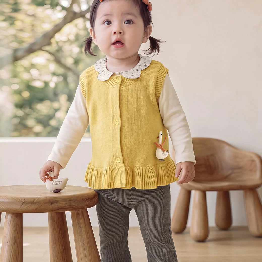 Little girl in a mustard knit vest standing by a wooden stool
