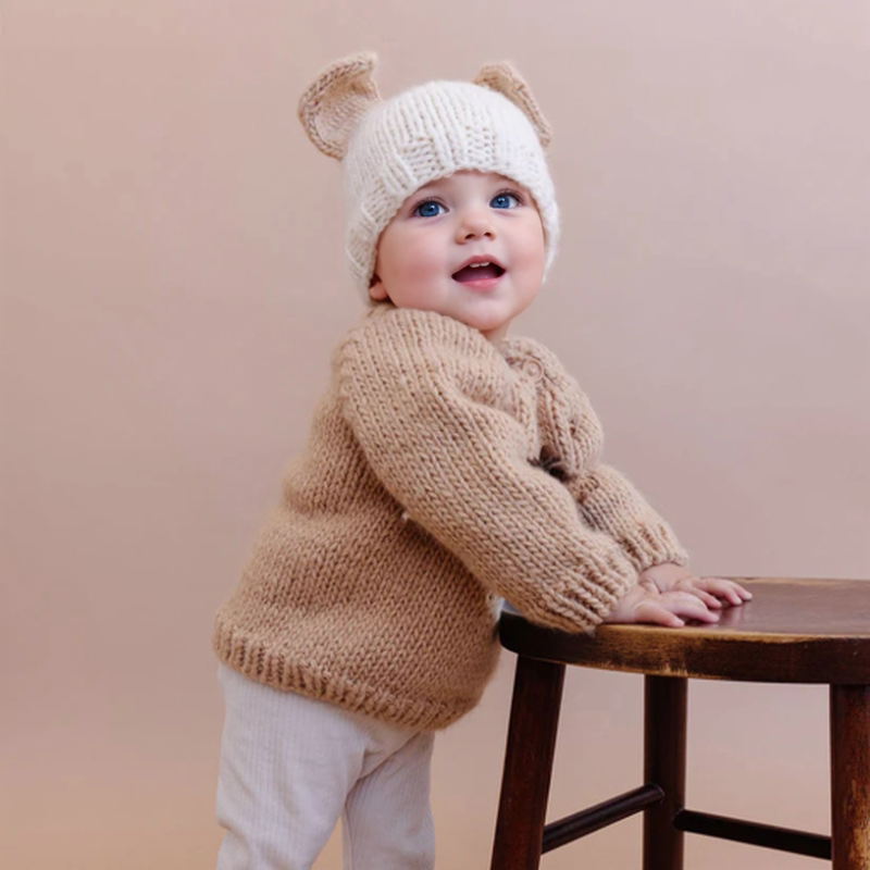 Smiling toddler in a camel chunky knit puppy sweater leaning on a wooden stool