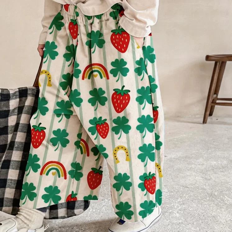 Kid in clover print summer pants with elastic waist, standing by a wooden stool