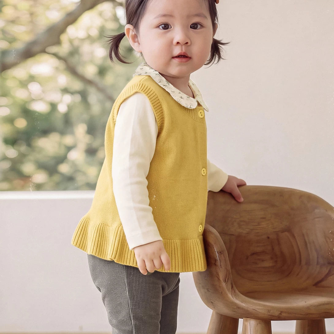 Baby girl in a mustard ruffle-hem sweater vest leaning on a wooden chair
