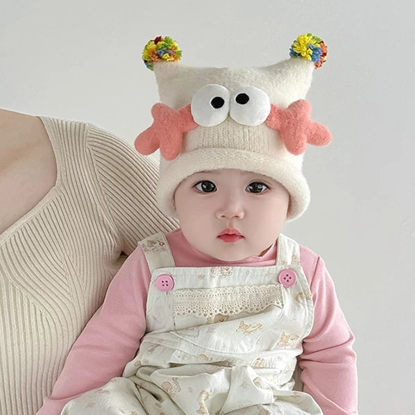 Baby in a cream googly-eye knit hat with rainbow pom-poms