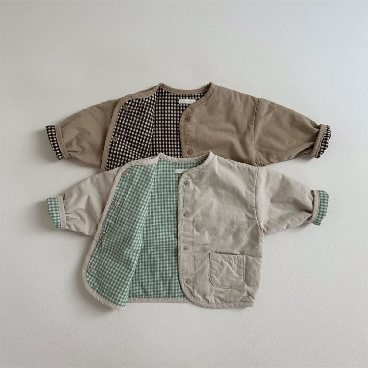Two reversible baby jackets in oat with sage gingham and mocha with brown check, flat lay
