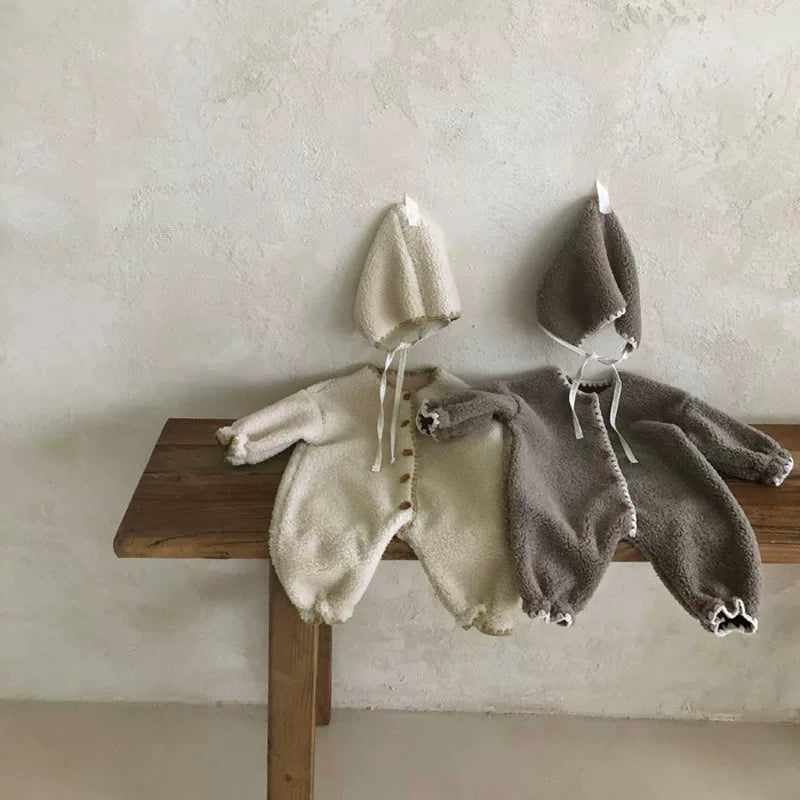 Plush baby romper sets with bonnets on a rustic wooden bench