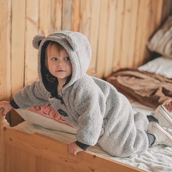 Baby climbing onto a bed in a cosy grey bear romper