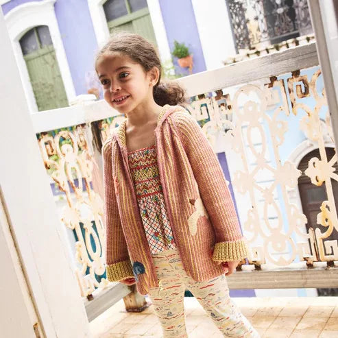 Girl in a rose knit hoodie cardigan by a white garden gate
