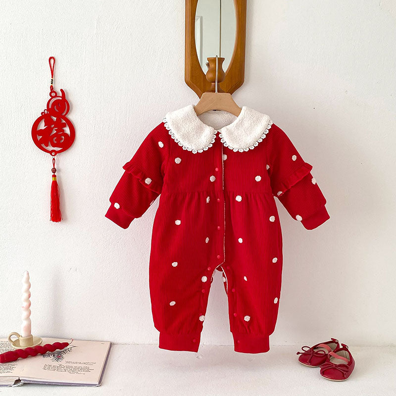 A vibrant red baby jumpsuit with white polka dots, featuring ruffle details on the sleeves and a white faux fur collar adorned with lace. The jumpsuit is displayed hanging on a wooden hanger in a minimalist setting, accompanied by decorative elements like a red tassel ornament, candles, an open book, and matching red baby shoes.