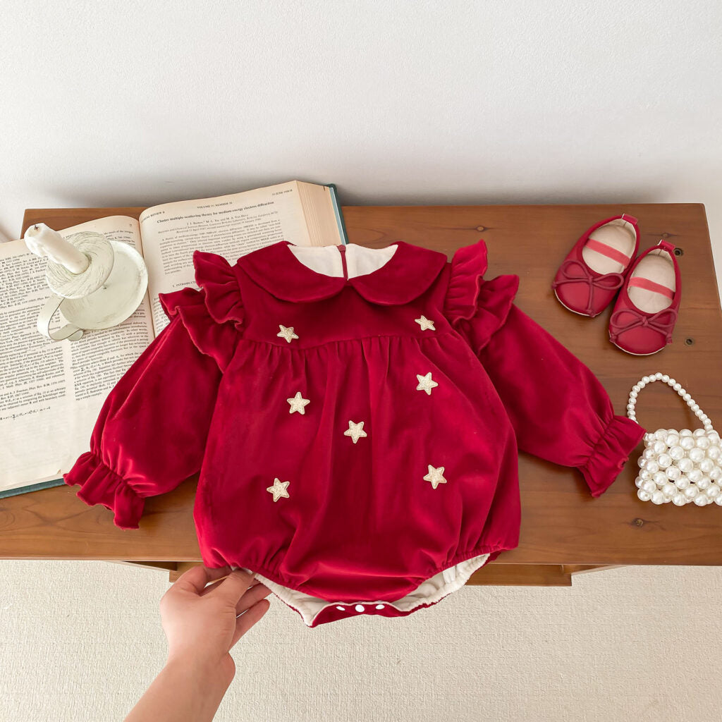 Elegant red velvet baby romper adorned with gold star accents, capturing a festive and whimsical look. The romper features a Peter Pan collar, ruffled shoulders, and puffed sleeves with elasticized cuffs for a charming silhouette. Paired with red baby shoes and a pearl handbag, it’s styled on a wooden table with open books and a decorative candle for a classic, cozy atmosphere.