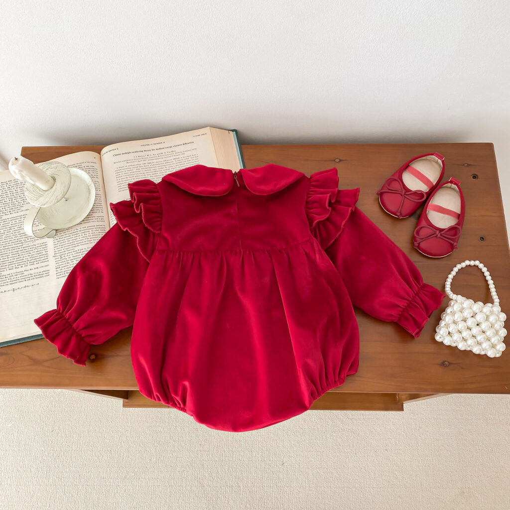 Back view of a red velvet baby romper showcasing a simple and elegant design. The romper features a concealed zipper closure, a Peter Pan collar, ruffled shoulders, and puffed sleeves with elasticized cuffs. Styled with matching red baby shoes and a pearl handbag on a wooden table alongside open books and a decorative candle for a cozy, classic setup.