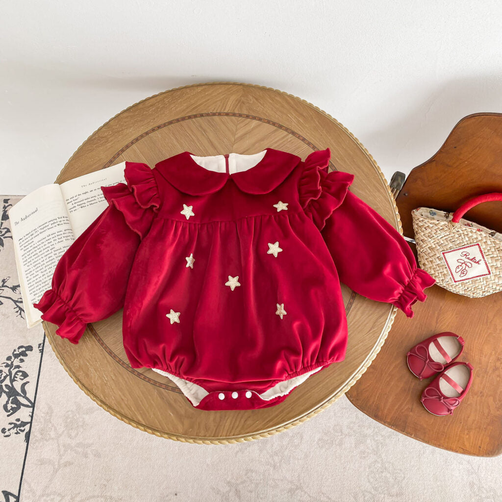 Twinkle Star Velvet Baby Romper displayed on a wooden round table. The romper features a Peter Pan collar, ruffled shoulders, and puffed sleeves with elastic cuffs. Embellished with star accents across the front, the romper has a snap-button closure for convenience. Styled with red baby shoes and a woven basket handbag featuring embroidered details, alongside an open book for an elegant and charming setup.