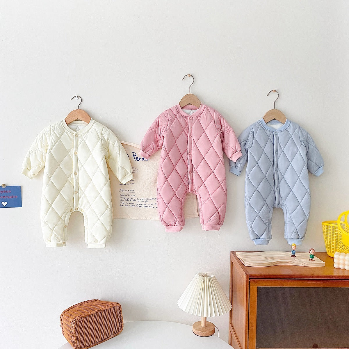 Three quilted baby jumpsuits with fleece lining hanging on wooden hangers against a light wall. The jumpsuits come in three colors: cream, pink, and light blue, each featuring diamond-pattern stitching, snap buttons, and long sleeves. The cozy design is perfect for keeping babies warm.