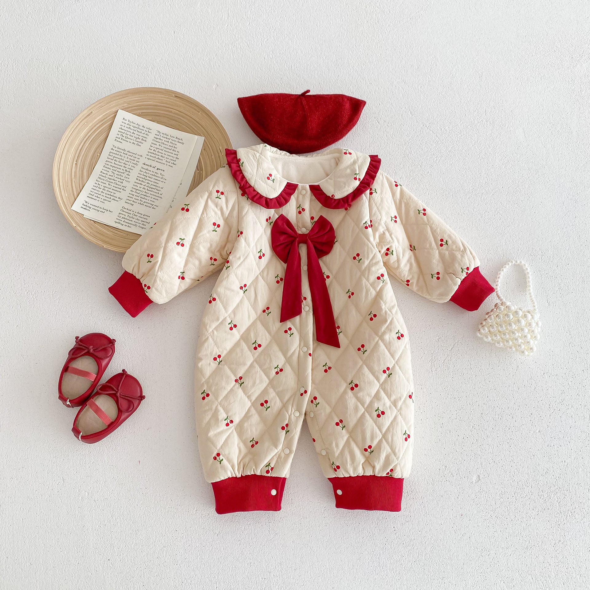 Adorable cream-colored baby jumpsuit with cherry prints, red cuffs, and a quilted texture. It features a ruffled red collar and a red bow on the chest, styled with a matching red beret, red baby shoes, and a pearl handbag for an elegant and festive look.