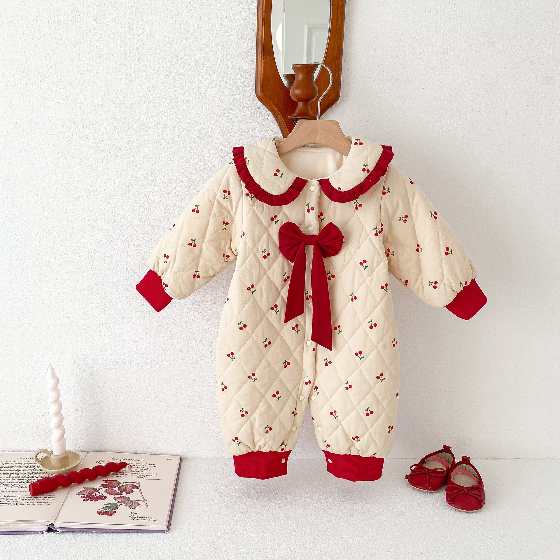An adorable cream-colored baby jumpsuit adorned with small cherry patterns, featuring a quilted design, red cuffs, a ruffled red collar, and a large red bow in the center. Displayed hanging on a wooden hanger with matching red baby shoes and decorative elements nearby, it showcases a cozy and festive winter look.