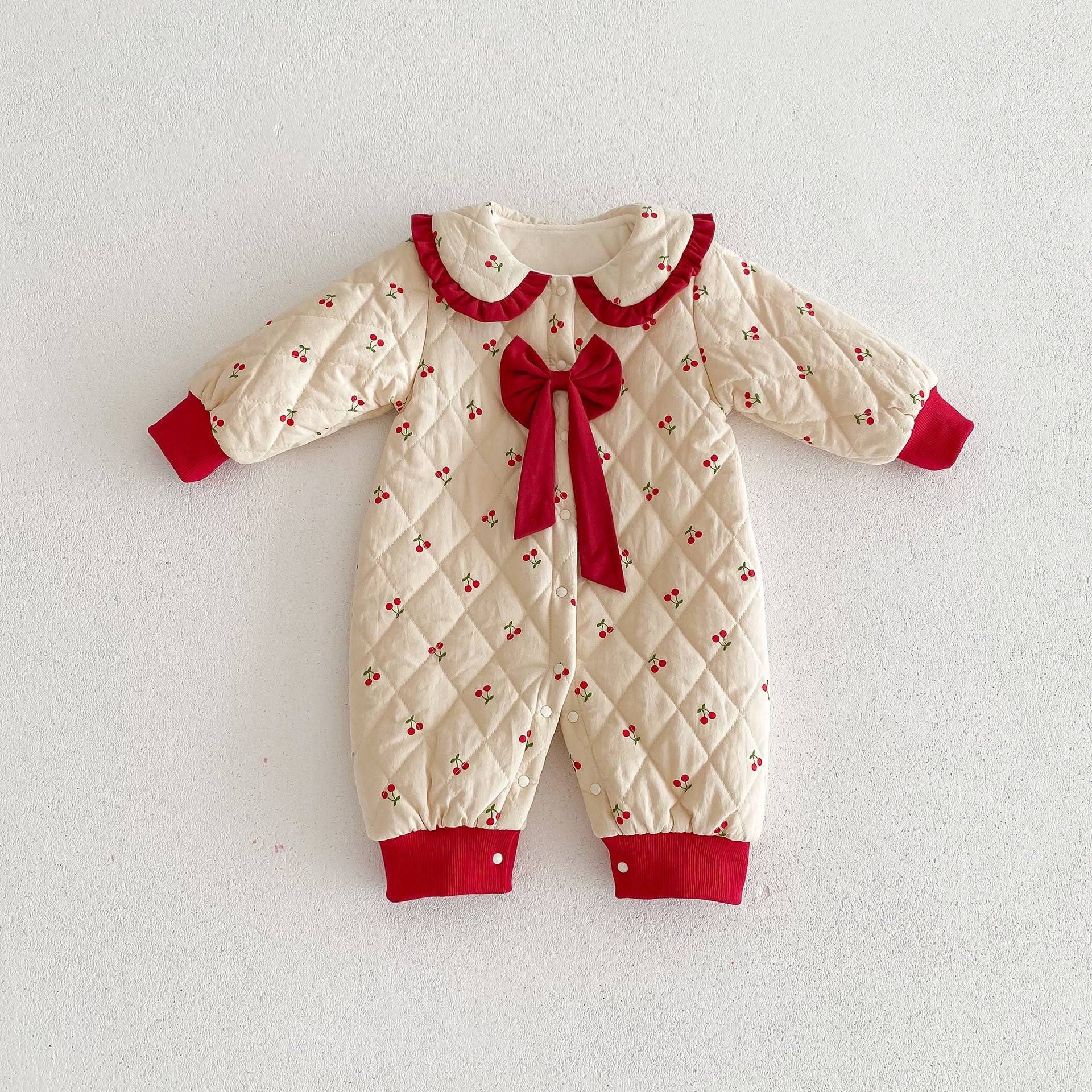 Adorable cream-colored baby jumpsuit designed with an all-over cherry print, featuring a cozy quilted pattern for warmth and comfort. This charming outfit is detailed with vibrant red cuffs, a large red bow accent on the chest, and a ruffled red collar that adds a festive and elegant touch. Perfect for special occasions or everyday wear during colder seasons