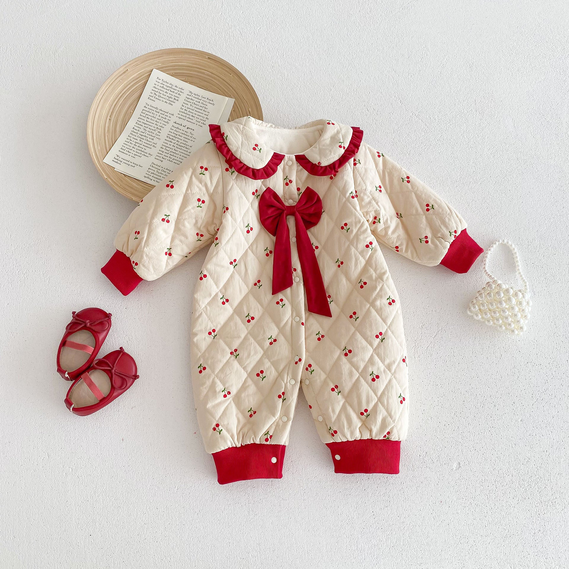 Cream-colored baby jumpsuit with small cherry patterns and a quilted design. It features red cuffs, a ruffled red collar, and a large red bow in the center. The jumpsuit is laid flat, styled with matching red baby shoes and a pearl handbag for a charming and festive look.