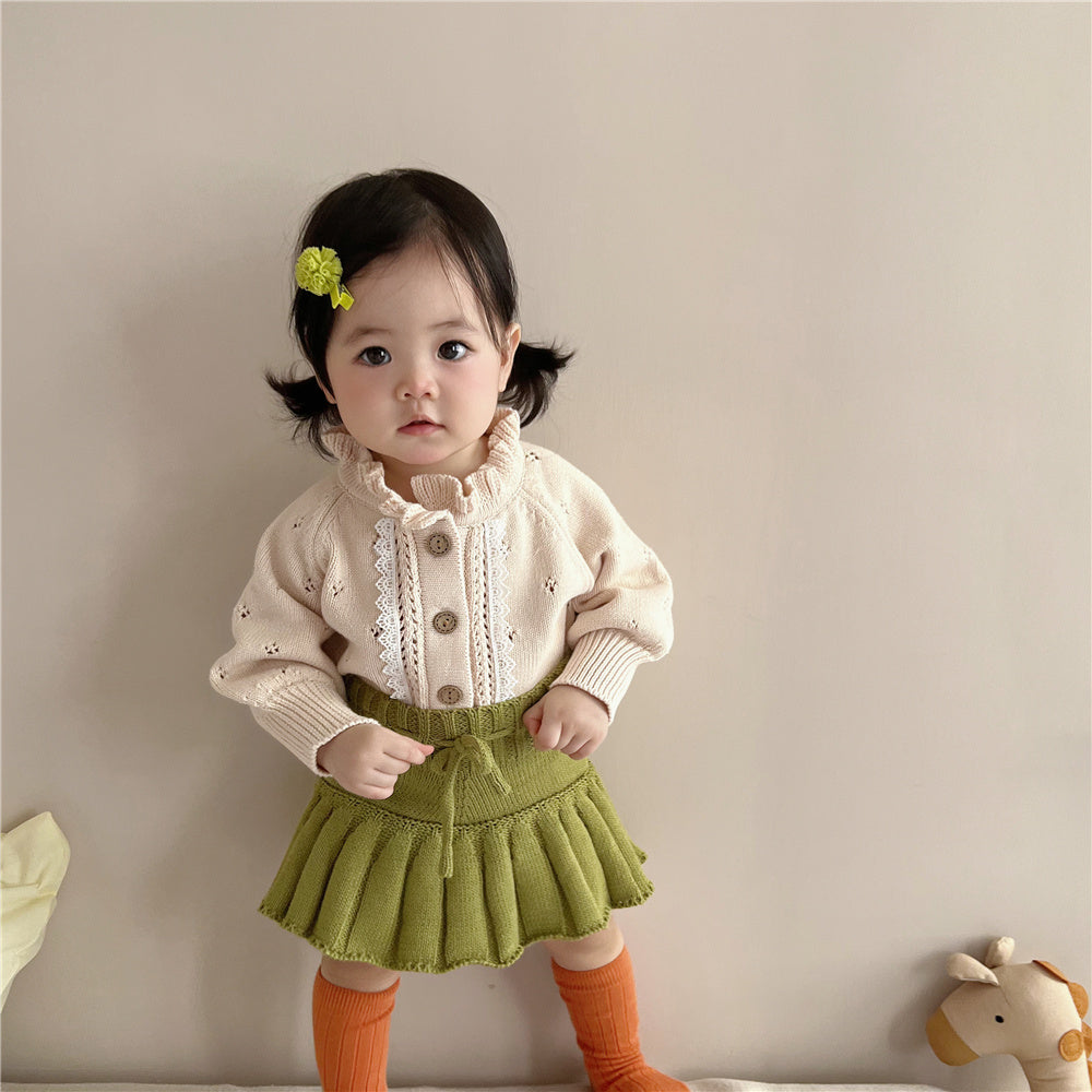 Baby girl in cream lace knit cardigan styled with green skirt and orange socks