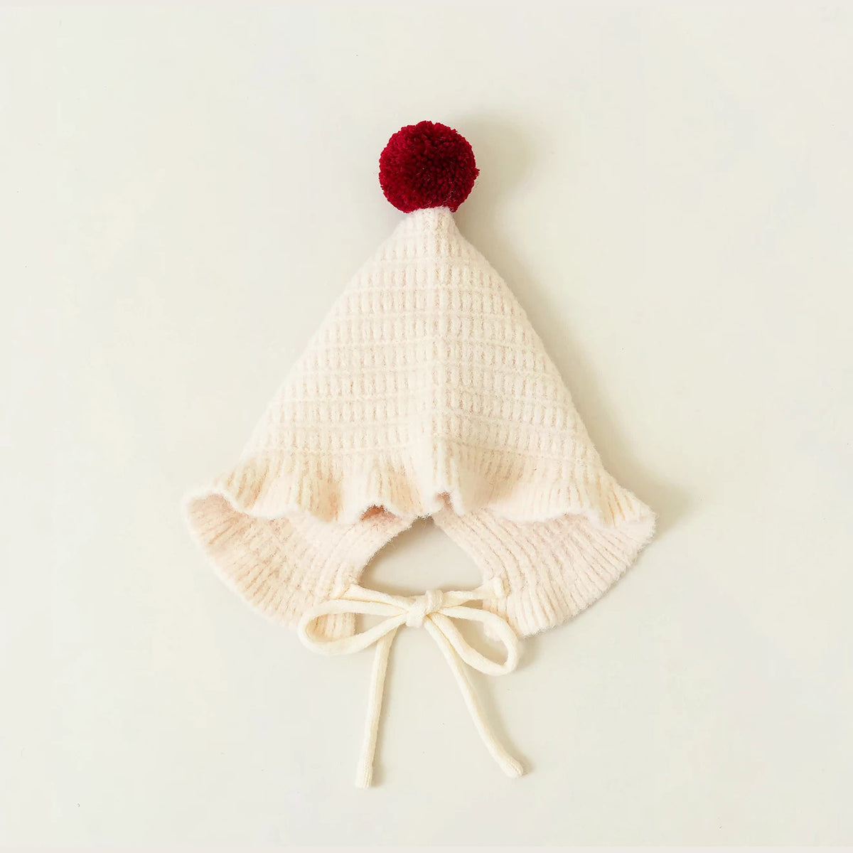 Cream pointed knit baby hat with a berry pom-pom on top, flat lay