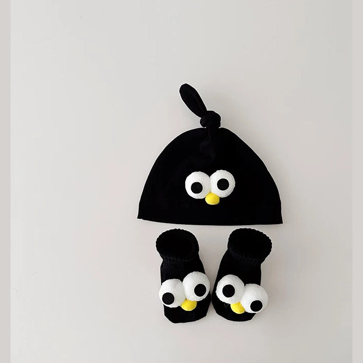 Black knot baby beanie and socks with googly eyes and a yellow beak, flat lay