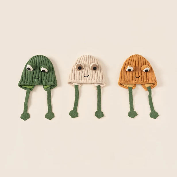 Three frog knit baby hats in green, oat and pumpkin with dangling frog-leg ties