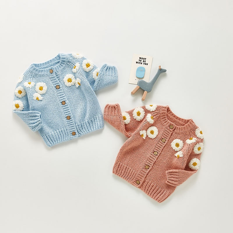 Cute baby cardigans in soft pastel colors, perfect for fall and spring. One cardigan is light blue, and the other is a warm pink, both featuring adorable embroidered white daisy flowers with yellow centers on the sleeves and shoulders. The button-up design with wooden buttons adds a vintage touch, while the floral pattern brings a cheerful, playful feel. Displayed alongside a small blue toy giraffe and a cute card