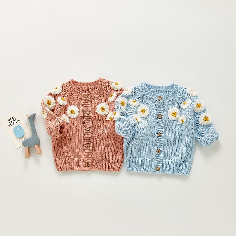 Set of two adorable baby cardigans in pastel colors, perfect for spring and fall. One cardigan is a warm pink and the other a soft blue, both featuring charming embroidered white daisies with yellow centers on the shoulders and sleeves. Each cardigan has a button-up front with wooden buttons for a rustic touch. Paired with a cute blue giraffe toy and a friendly card,
