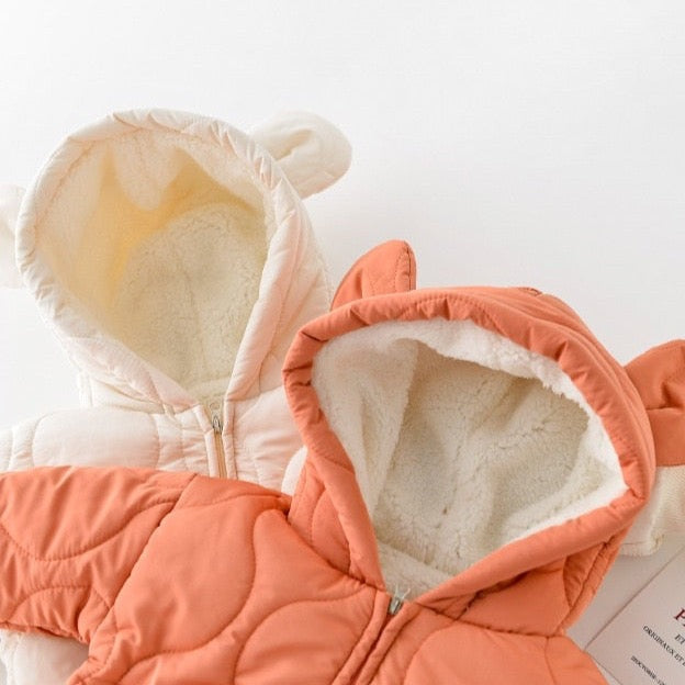 Close-up view of two quilted baby jumpsuits, one in cream and the other in burnt orange. Each jumpsuit features a cozy, fleece-lined hood with cute bear ears, adding a charming touch to the warm and soft design, perfect for keeping babies snug during chilly weather.
