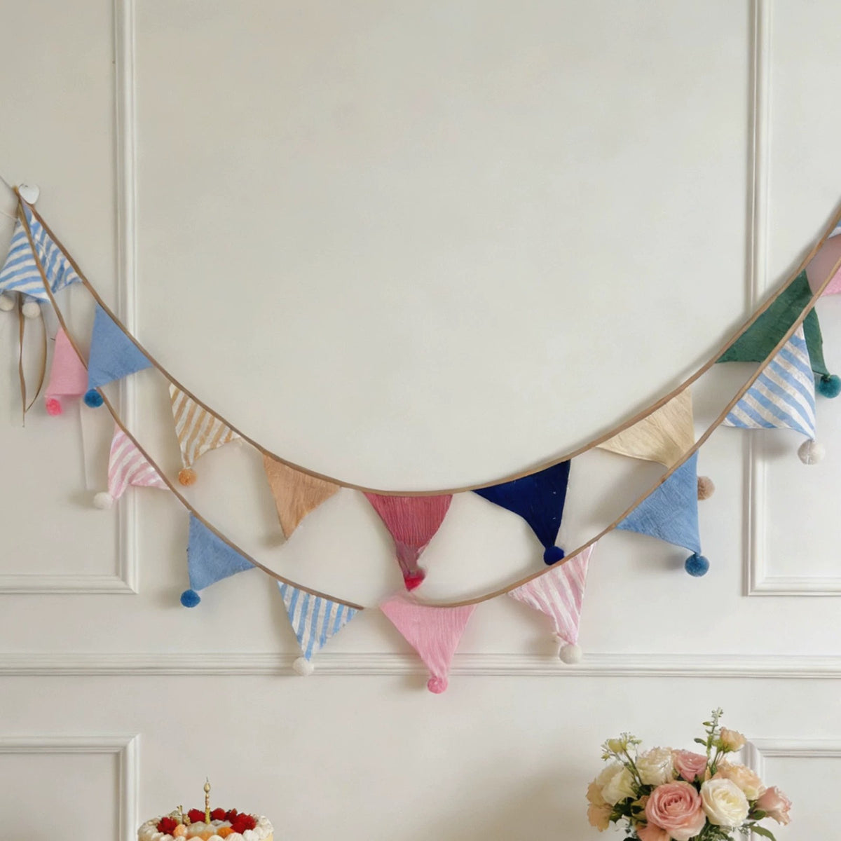 Muslin pom-pom bunting draped across a white panelled wall above a birthday cake