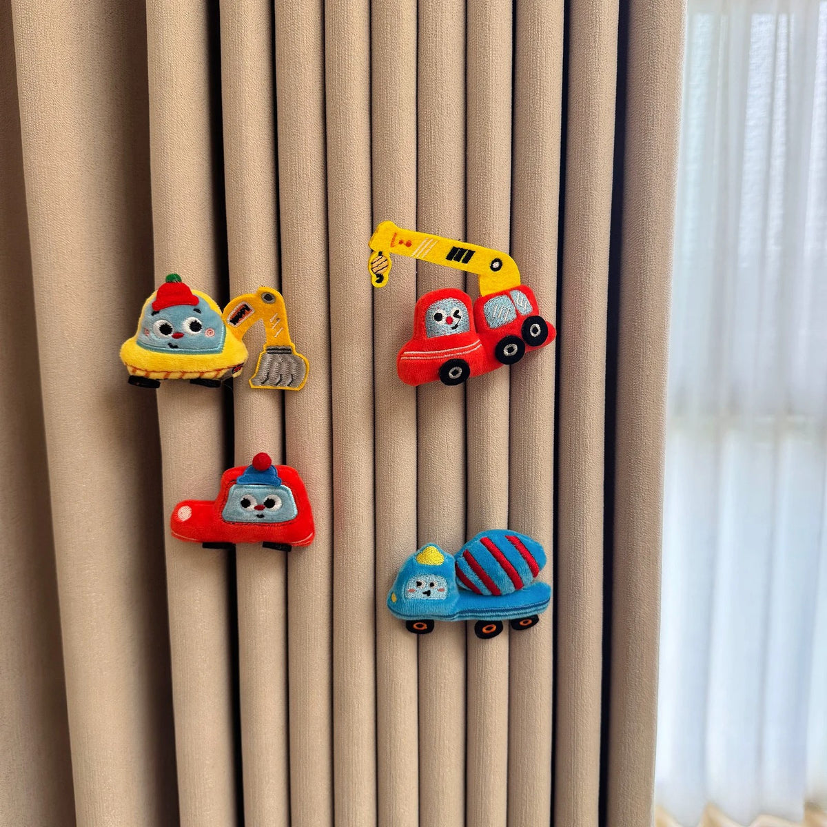 Four plush vehicle pins – excavator, crane, police car and mixer truck – on a beige curtain