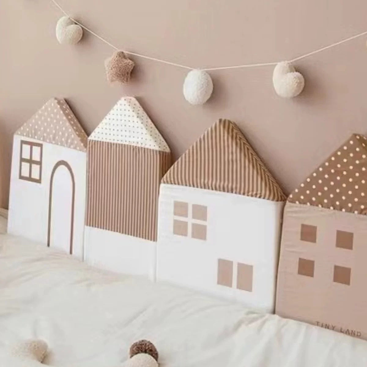 Mocha little house cushions lined up along a bed below a pom-pom garland
