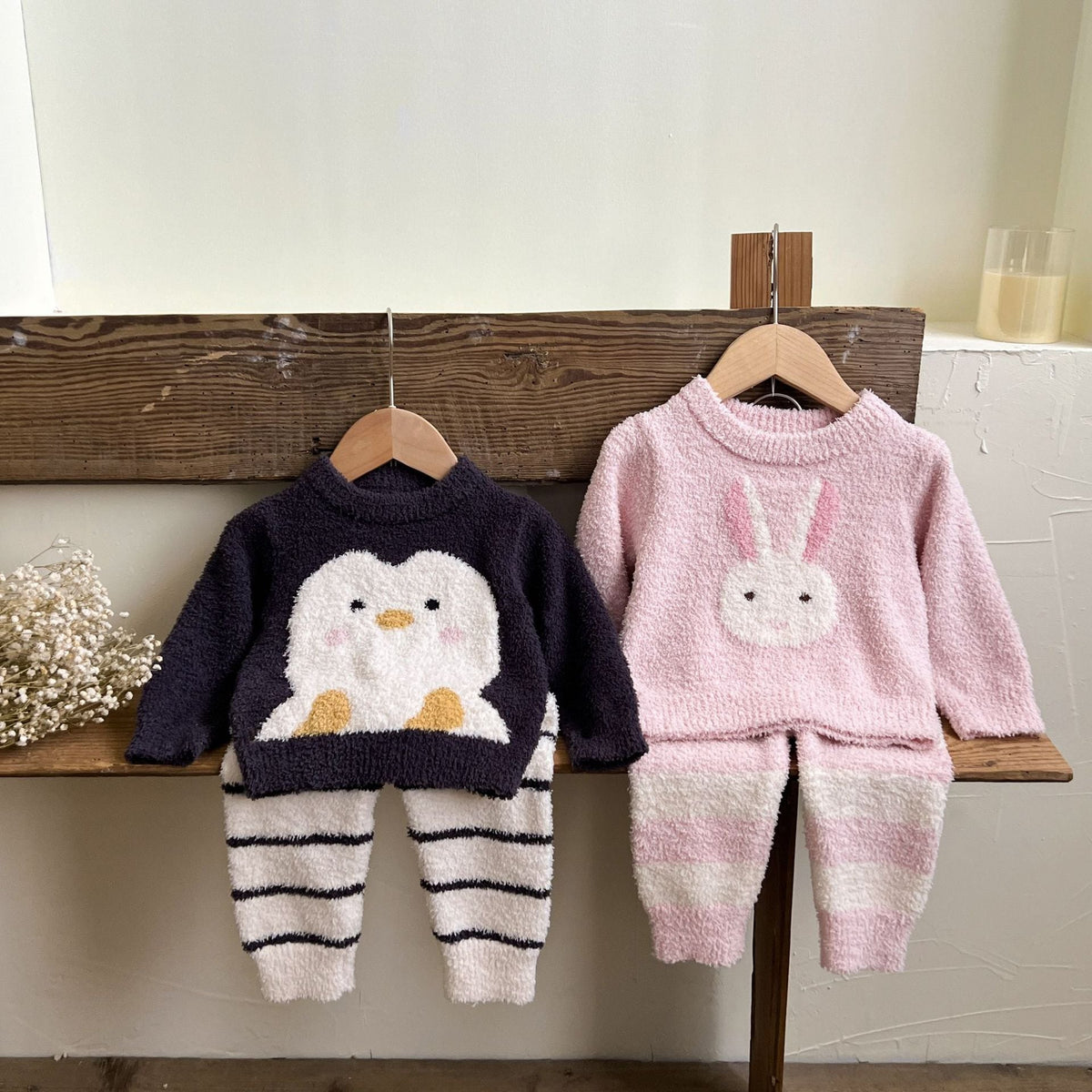Penguin and bunny knit sets on hangers against a wooden bench