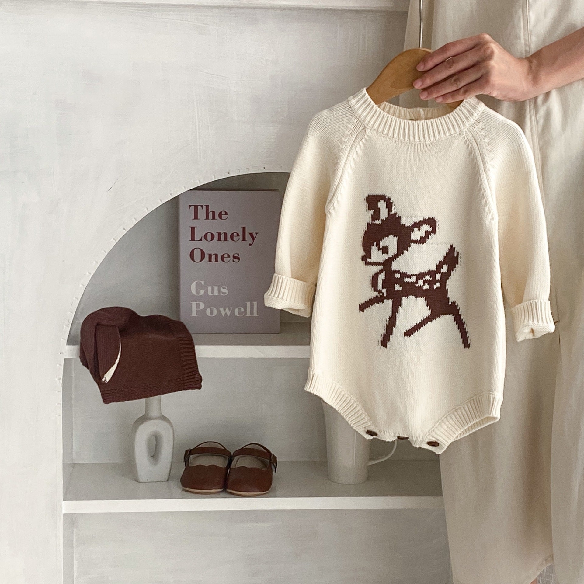 Stylish baby outfit display featuring a cream-colored knit romper with a cute brown deer pattern, ideal for fall and winter baby fashion. The romper is held on a wooden hanger by a hand, with a shelf backdrop displaying matching brown accessories, including a cozy hat with ear details and a pair of classic baby shoes. Also styled with a minimalist decor touch, featuring the book 'The Lonely Ones' by Gus Powell, adding a literary vibe. Perfect for a modern, aesthetically pleasing baby clothing photoshoot