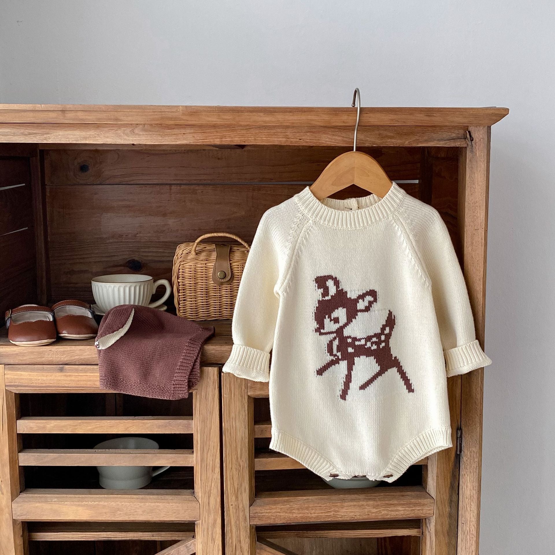 Alt text: "Charming baby outfit arrangement with a cream-colored knit romper featuring an adorable brown deer design, perfect for a cozy winter look. The romper hangs on a wooden hanger against a rustic wooden cabinet, complemented by matching accessories including a brown beanie hat with ear details and a pair of classic baby shoes. The setting includes a woven basket and a white ceramic cup, adding a warm, vintage feel to the display.