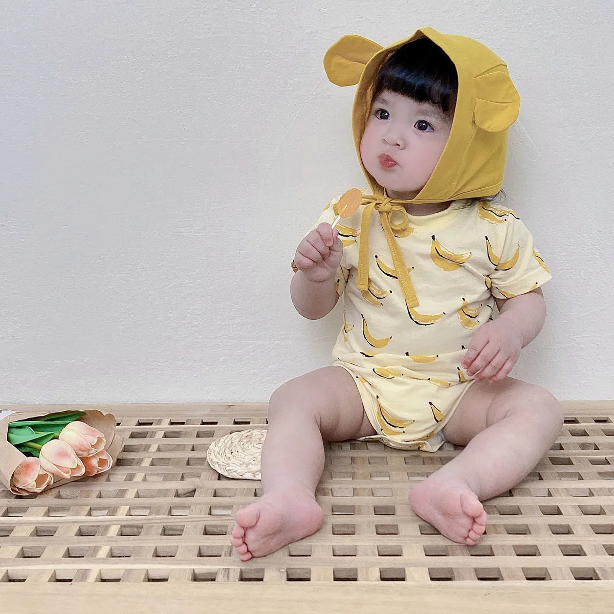 Baby girl in banana print bodysuit and bear bonnet sitting with lollipop