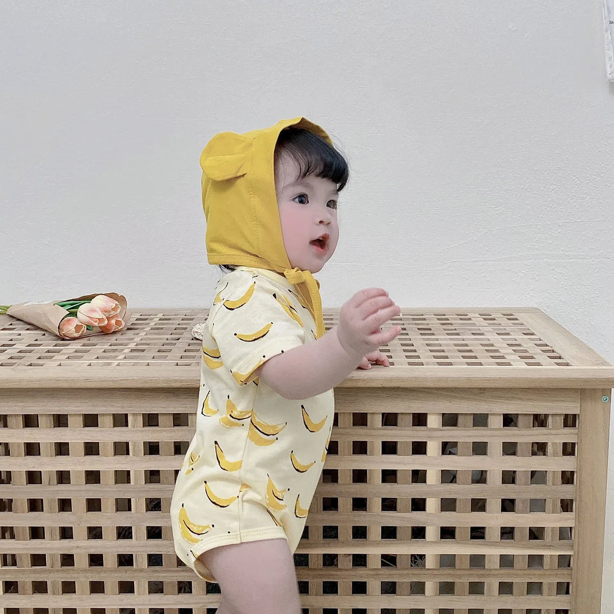 Baby wearing banana bodysuit + bonnet, side profile, reaching toward wooden crate