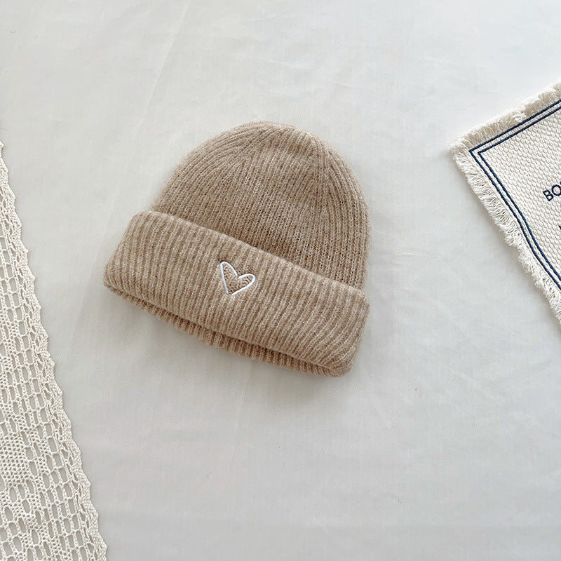 Soft beige knitted beanie with a ribbed texture and an embroidered heart detail on the cuff. This cozy winter hat combines warmth and minimalist charm, making it a stylish addition to any cold-weather outfit. Displayed on a neutral background with lace and fabric accents, ideal for showcasing in e-commerce listings or seasonal accessory catalogs.