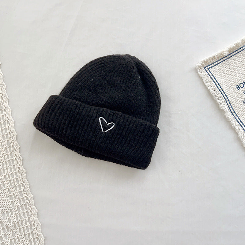 Classic black knitted beanie with a ribbed texture and a small white embroidered heart detail on the cuff. This minimalist winter hat adds a subtle, stylish accent to any cold-weather outfit. Displayed on a neutral background with lace and fabric accents, perfect for e-commerce listings or seasonal fashion catalogs.