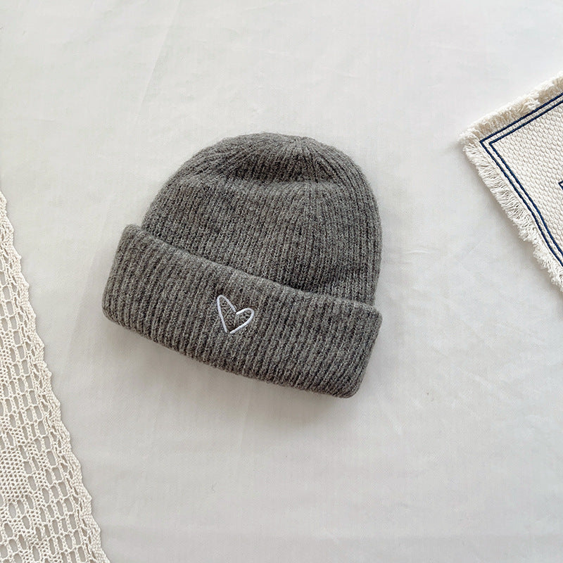 Gray knitted beanie with a ribbed texture and a small white embroidered heart on the cuff. This cozy winter hat is designed to provide warmth and add a subtle touch of charm to any cold-weather outfit. Displayed on a minimalist background with lace and fabric accents, ideal for showcasing in e-commerce listings or seasonal fashion catalogs.