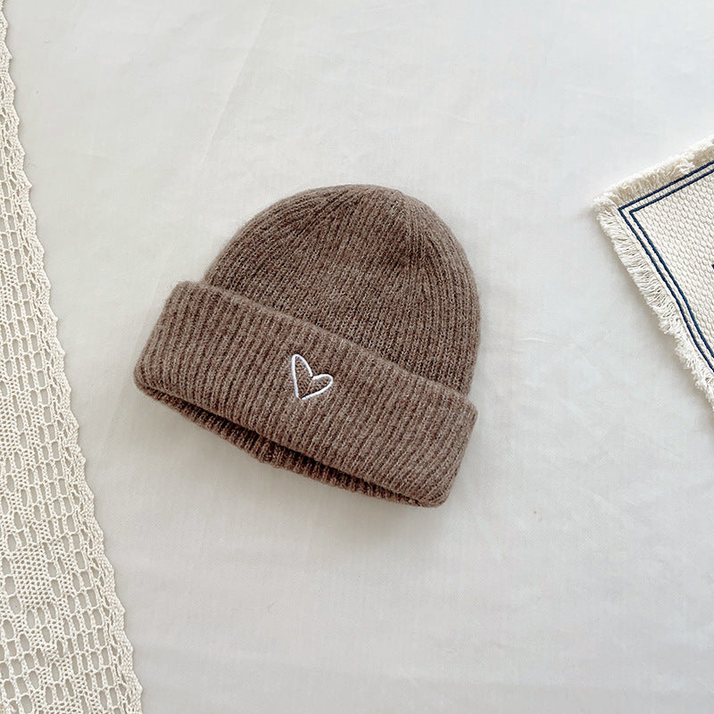 Warm brown knitted beanie with a ribbed texture and a small white embroidered heart on the cuff. This cozy hat adds a touch of charm to winter outfits, perfect for cold-weather wear. Displayed on a minimalist background with lace and fabric details, ideal for product listings in an online store or seasonal fashion catalog.