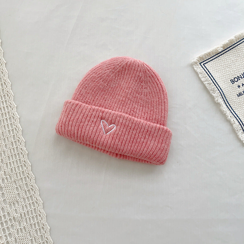 Soft pink knitted beanie with a ribbed texture and a small white embroidered heart on the cuff. This charming hat is perfect for a cozy, warm look, especially suited for cold weather. Displayed on a light background with lace and fabric details, ideal for product listings in a seasonal fashion collection or online boutique.