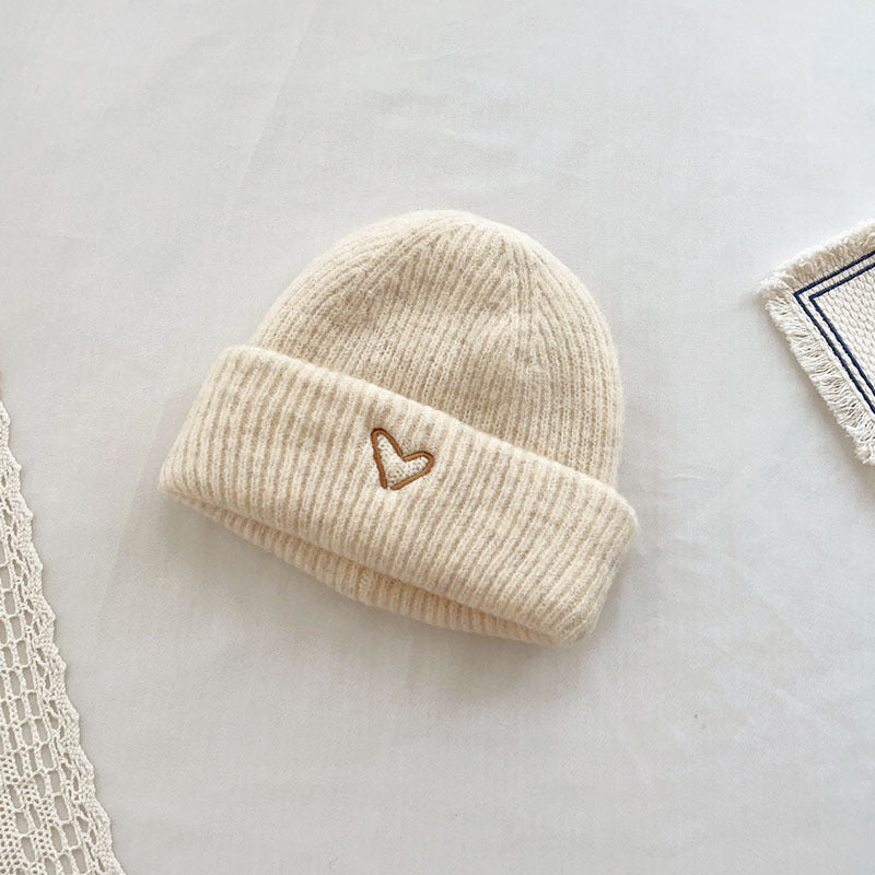 Soft cream-colored knitted beanie with a cozy, ribbed texture and an embroidered heart design on the cuff. This minimalist, stylish hat is perfect for keeping warm in cooler weather while adding a touch of charm to any outfit. Displayed on a neutral background with delicate lace and fabric details, making it ideal for showcasing in e-commerce listings or winter accessory catalogs.