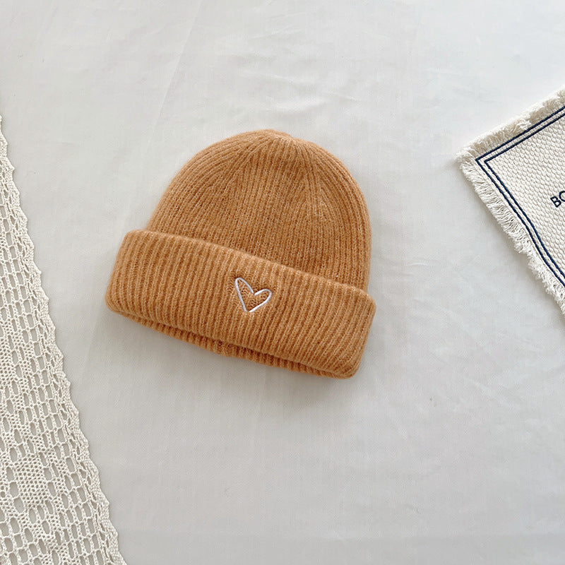 Warm tan-colored knitted beanie with a ribbed texture and a small embroidered heart detail on the cuff. This cozy hat adds a charming touch to any winter outfit, perfect for keeping warm in style. Displayed on a neutral background with lace and fabric accents, making it ideal for showcasing in e-commerce listings or winter accessory catalogs.