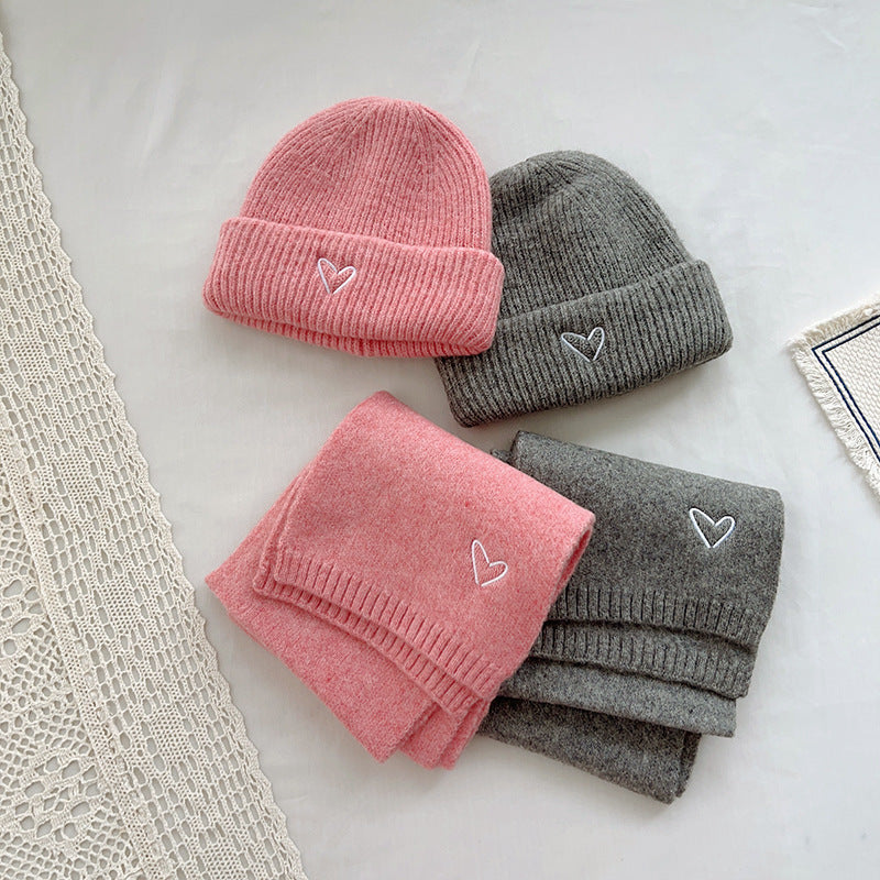 Stylish winter accessory set featuring matching knitted beanies and scarves in pink and gray, each adorned with a small embroidered heart for a subtle, charming detail. The ribbed texture of the beanies and the soft, cozy fabric of the scarves make them perfect for cold weather, adding both warmth and a pop of color to any outfit. Displayed on a minimal background with lace and fabric elements, this set is ideal for showcasing in e-commerce listings or seasonal fashion catalogs.