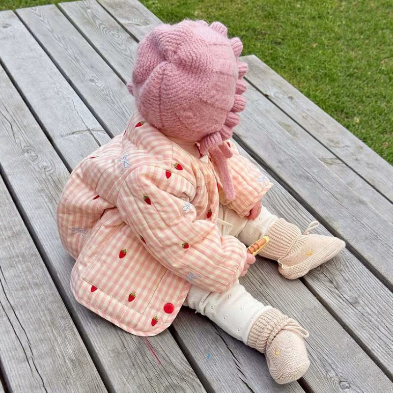 Toddler in a blush pink strawberry gingham jacket sitting on a wooden deck