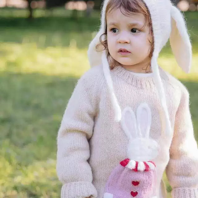 Little girl in a cream knit sweater with a pink bunny motif, standing in a park