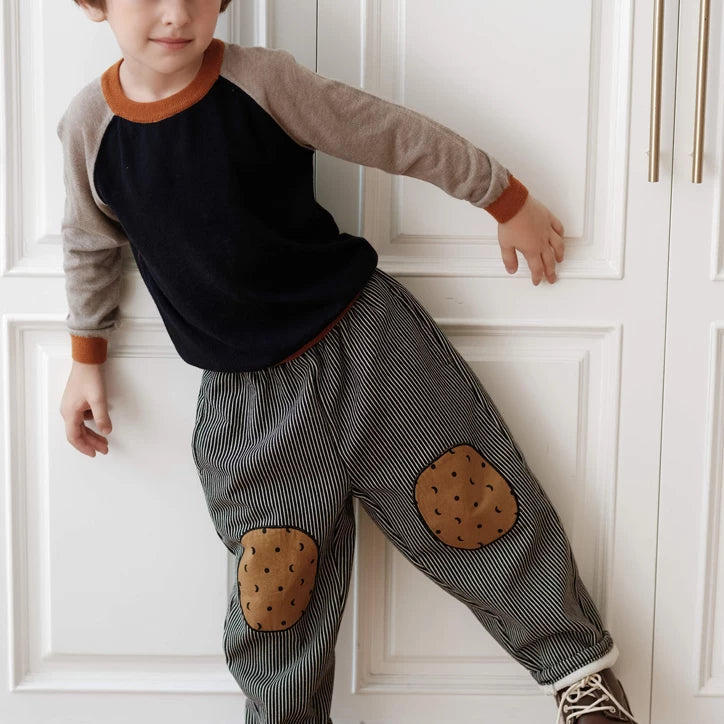 Child leaning on a white door in striped kids pants with brown knee patches