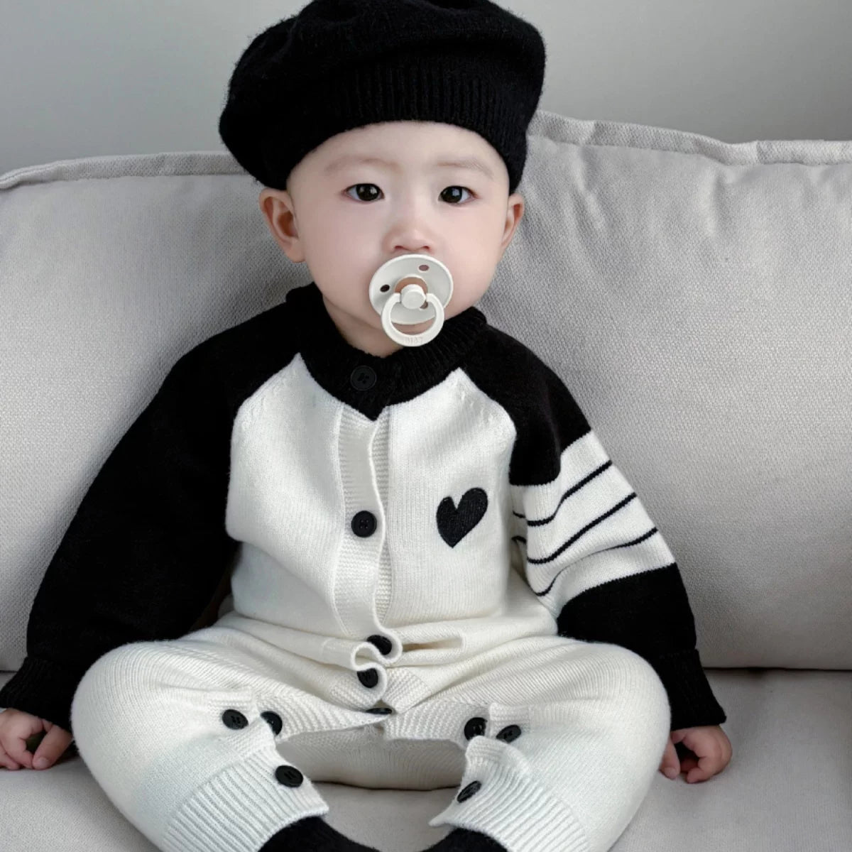 Baby in a black beret wearing the cream and black heart knit romper on a sofa