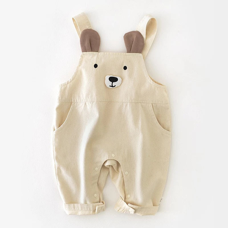 Beige cotton baby overalls with a bear face and brown ears on the straps, flat lay
