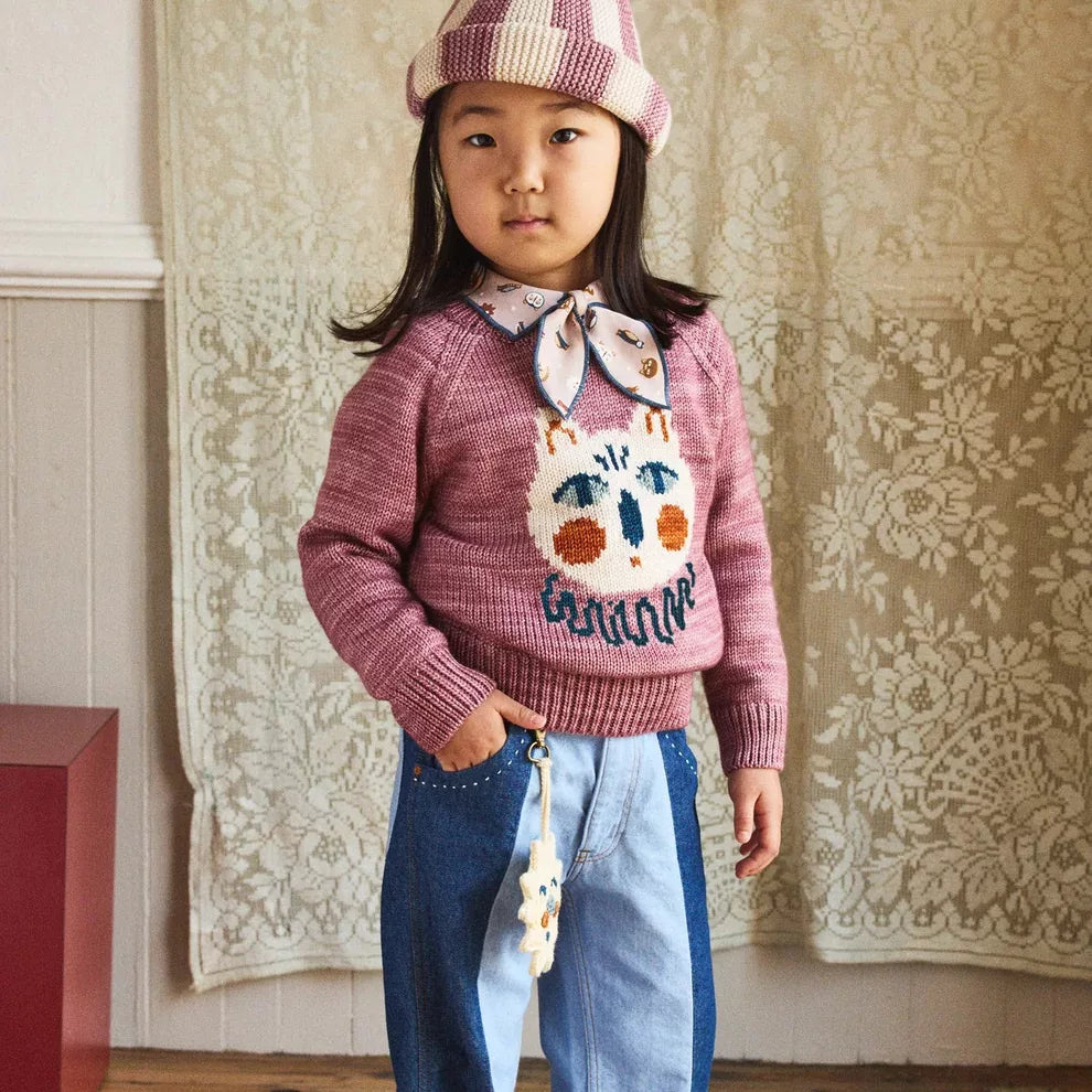 Girl in a berry knit sweater with a cream cat face, wide-leg jeans and a pompom hat